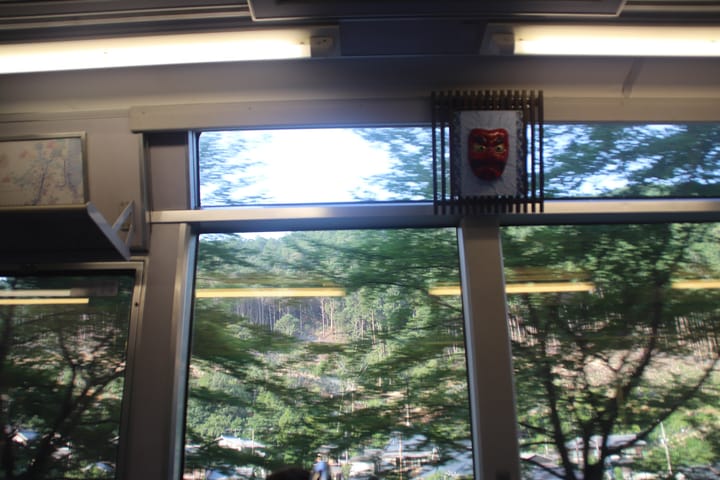 view inside a train looking out the window to trees, and a red mask on the wall of the train, thumbnail